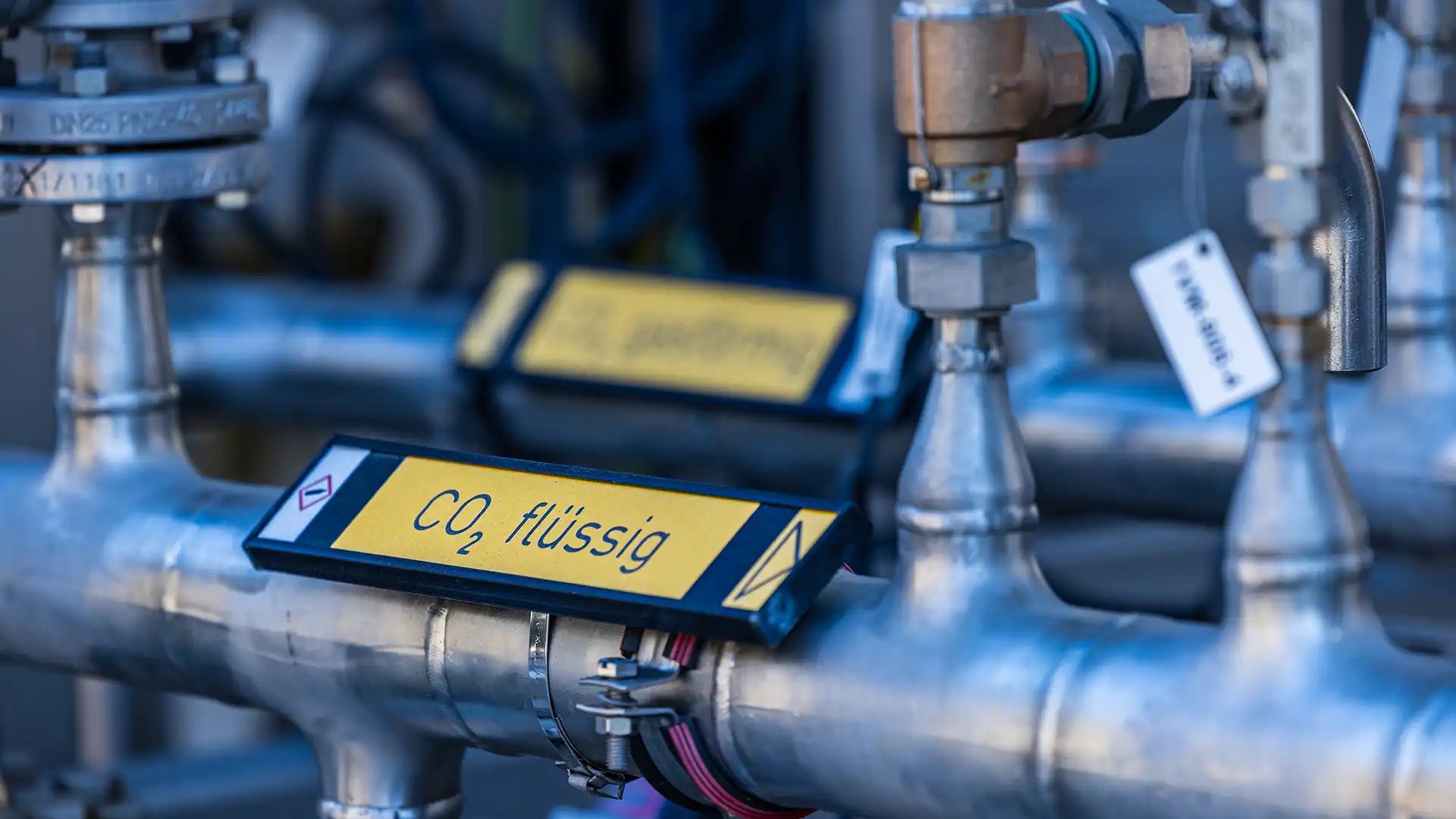 Close-up of industrial metal pipes inside a CO₂ recovery facility with a yellow label reading “Liquid CO₂”.